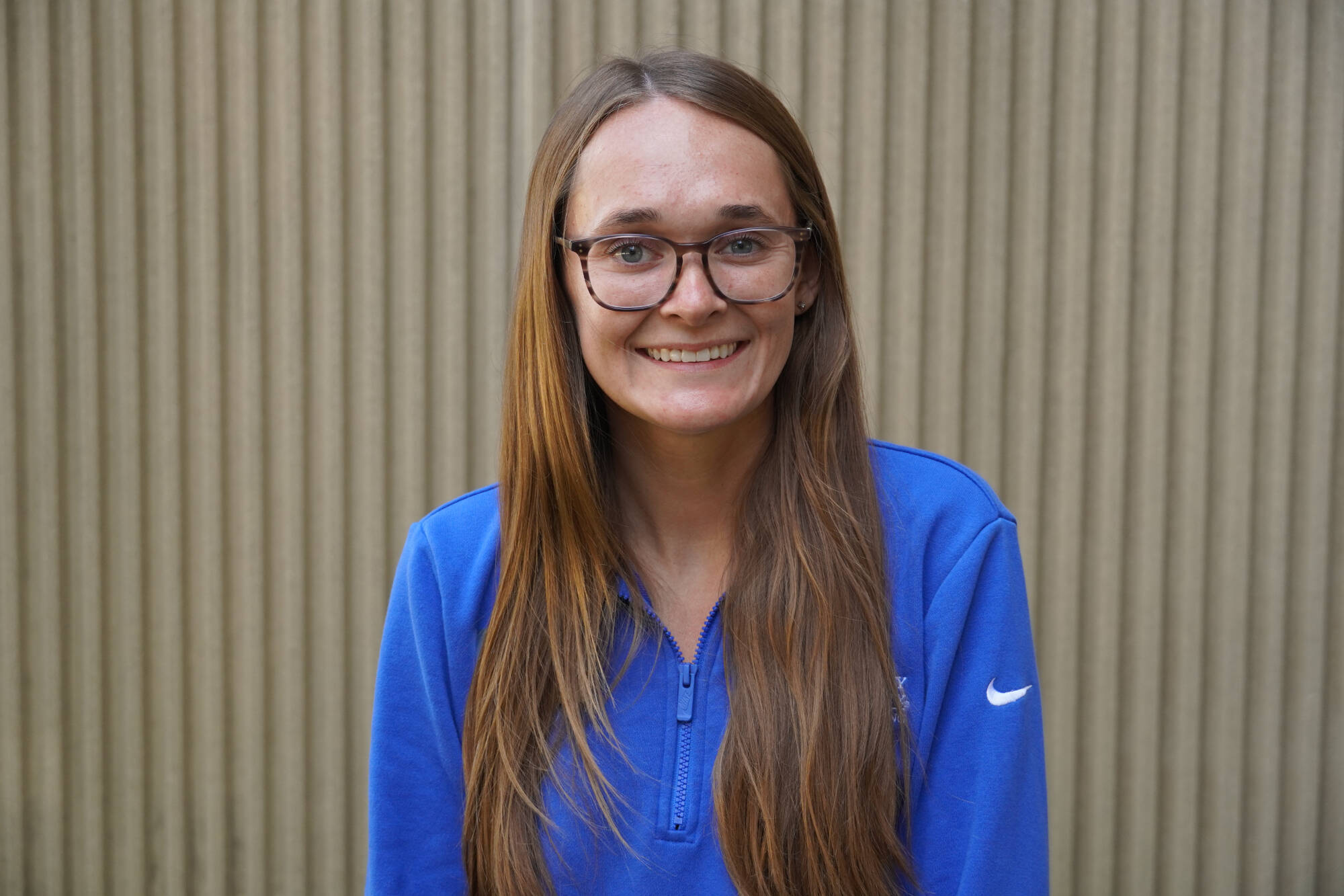 Image of Emily Zeliasko wearing a blue quarter zip in front of a textured wall as she is smiling.